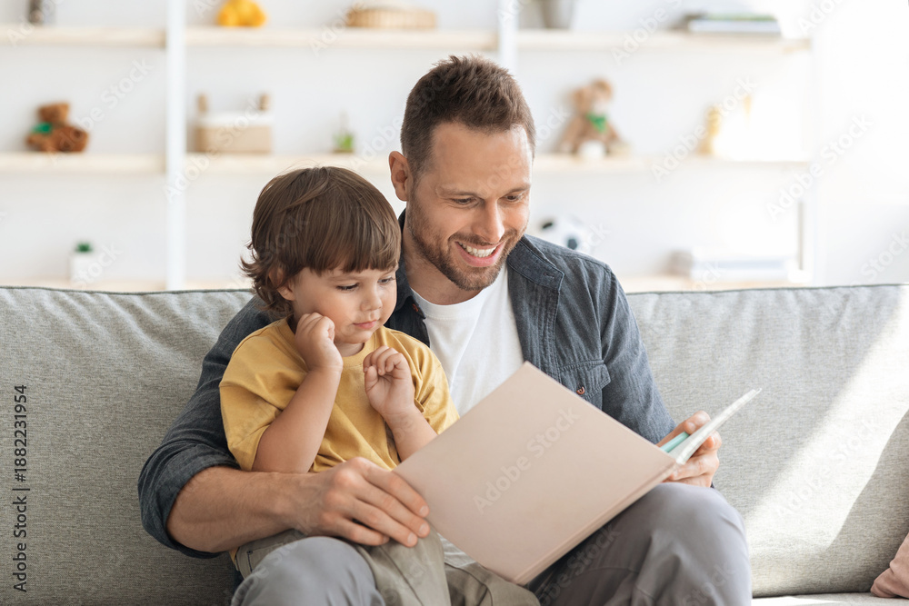 Fototapeta premium Story time. Loving father reading interesting fairy tales book to his little son, interested adorable boy enjoying time with his daddy, sitting together on sofa at home