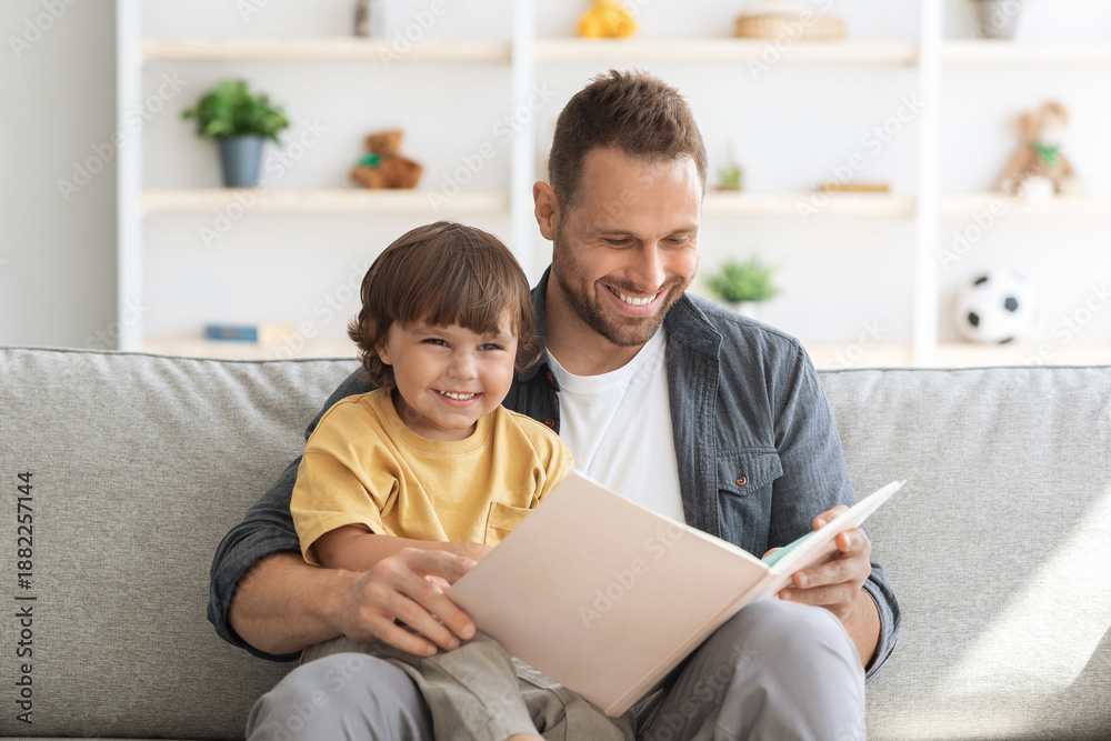 Fototapeta premium Storytime. Young loving father reading fairy tales to his cute little son, sitting together with big child book on sofa at home and smiling