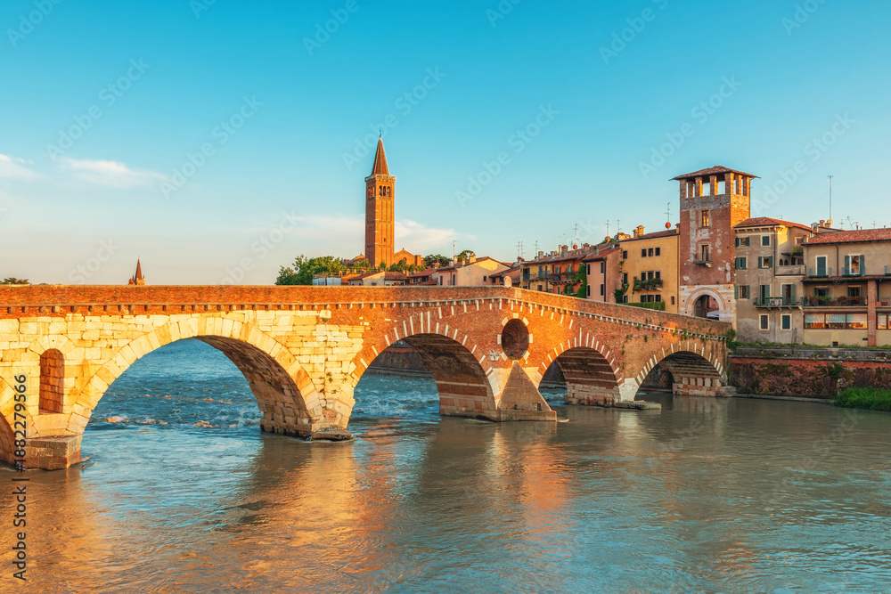 Fototapeta premium Historic stone Ponte Pietra bridge in Verona old town crossing Adige River with historic buildings and church tower at sunrise, Veneto, Italy. Skyline of Italian city. Travel destination in Europe