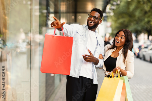 Wow, Big Sale. Portrait of emotional cheerful African American couple walking near shopping centre, looking at showcase, guy pointing finger at mall window. Seasonal Discount, Great Special Offer