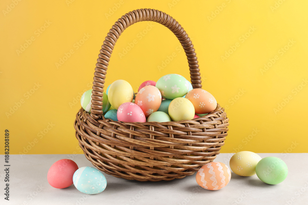 Fototapeta premium Beautiful Easter eggs in basket on white table against yellow background
