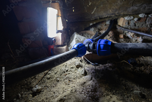 Fotografia Water pipes connecting with blue compression fittings on a dirt floor in a dark basement or crawl space, illuminated by a work light, highlighting household plumbing maintenance