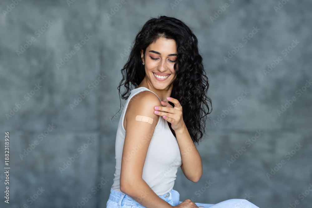 Fototapeta premium Coronavirus vaccination concept. Young Caucasian brunette woman showing adhesive bandage on her arm, getting vaccinated against covid-19 on grey background. Immunization for viral disease