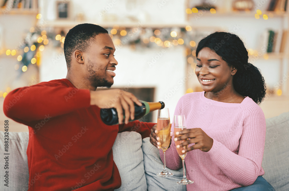 Obraz premium A man pours sparkling drink into glasses held by a woman as they sit on a couch. The room is decorated for a celebration with soft lights. They are enjoying a fun moment together.