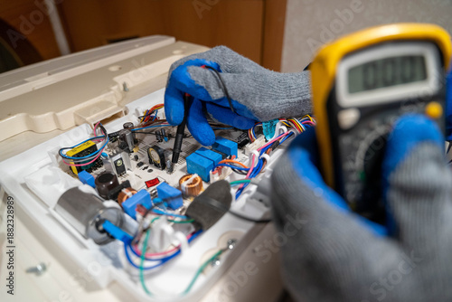 Technician wearing blue gloves is using a multimeter to test electronic components on a circuit board inside a workspace with visible tools and equipment