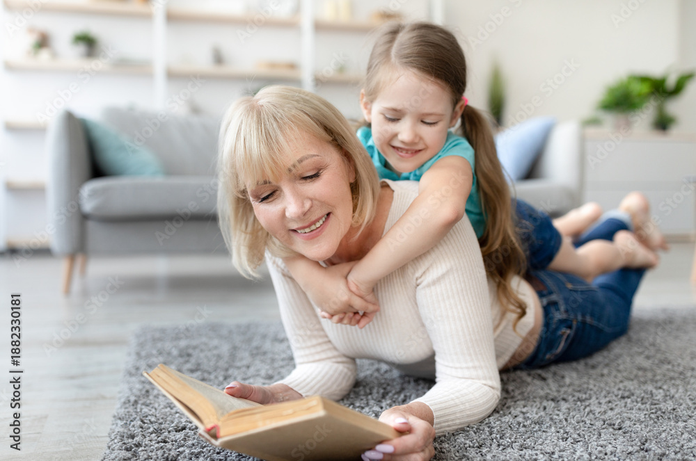 Obraz premium Loving grandmother teaching granddaughter holding book lying on floor carpet, kid girl embracing grandma babysitter, resting on her back, nanny reading fairytale story to cute preschool grandchild