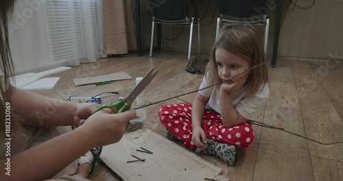 A young mother teaches her adorable daughter how to make crafts out of cardboard and twigs. The family sits together on the floor at home, working on a fun and educational family project.