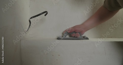 Close-up of a construction worker's hand sanding a plastered surface with a sanding block, creating dust, and then checking the smoothness of the wall with their palm and fingers.
