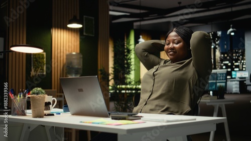African american woman reclining in a chair in a relaxed pose at midnight, daydreaming during work break. Fulfilled and self assured, reflecting on career success and achievements. Camera A.