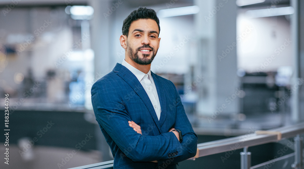 Fototapeta premium A man in a blue suit stands with arms crossed in a bright office. The scene shows an open work area with desks and modern equipment in the background.