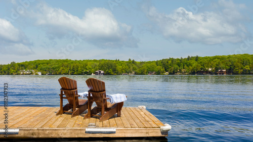 Two wooden Adirondack chairs on a private dock with folded towels overlook a calm Muskoka lake in Ontario, Canada. A pine-lined shore and clear summer sky evoke relaxed cottage life.