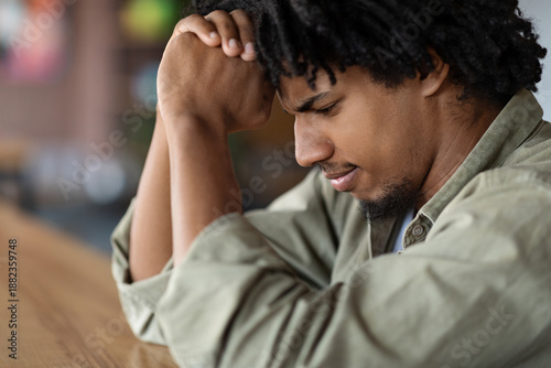 Фотография Sad serious concentrated young african american curly male praying or thinking about solving problem at table in cafe interior, profile, close up, cropped