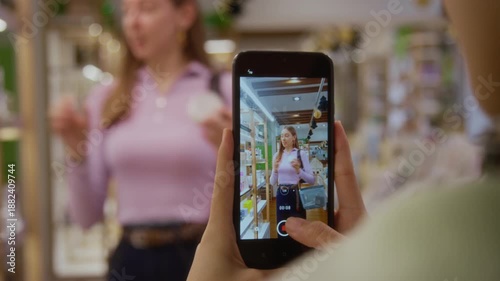 Rack focus from smartphone screen of female influencer describing nourishing hair balm on camera promoting skincare product while shooting video with friend for digital marketing at cosmetics store