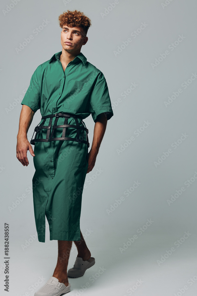 Fototapeta premium Young man with curly hair wearing green shirt dress and black belt walking barefoot on light background. Fashion model posing in casual outfit with relaxed expression and modern style.