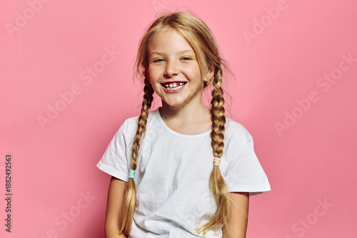Quadro su tela Smiling young girl with blonde hair in braids wearing a white t-shirt stands against a pink background