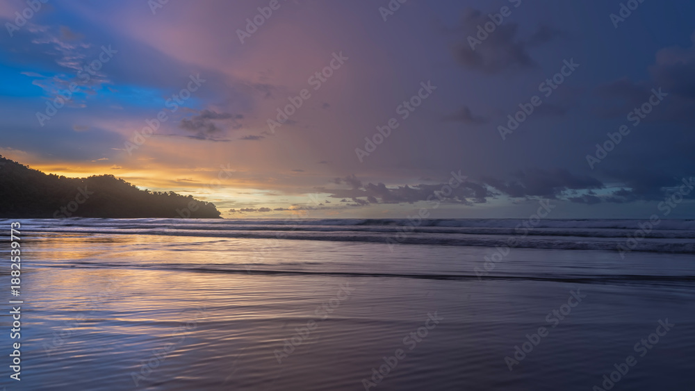 Fototapeta premium Beautiful colorful sunset over the ocean. The blue sky is highlighted with gold at the horizon. Purple and pink clouds. Waves are spreading across the beach. Reflection. A hill in the distance. 
