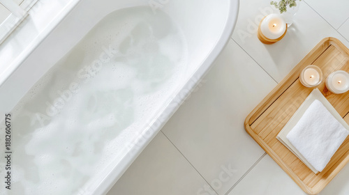 Bathtub filled with water and foam bubbles next to wooden tray with candles and towel creating calm and relaxing atmosphere