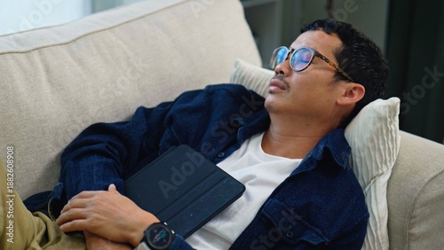 Foto Asian Man Falling Asleep on Sofa While Holding Tablet on His Chest