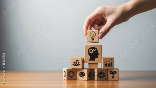 Hand building a tower with wooden blocks featuring business icons on a wooden desk with a gray background.
