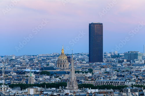 Distant cityscape view of Paris with the Montparnasse Tower and the golden dome of Les Invalides standing out against pastel evening skies, surrounded by dense urban rooftops.