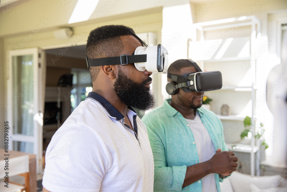 Fototapeta premium African american male friends testing vr headsets standing in living room with sliding glass doors