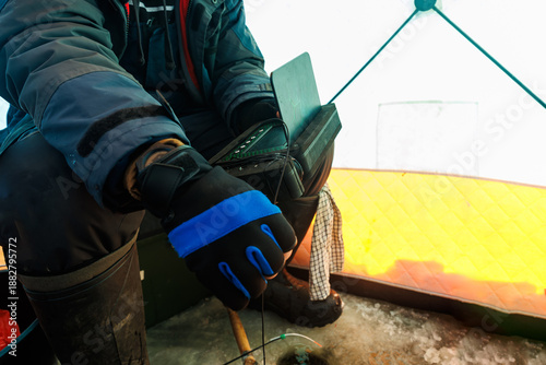 Fotografie Inside a quilted ice shelter, an angler in winter gear uses a compact sonar and holds a transducer over a drilled hole, with hand tools nearby in bright daylight winter light