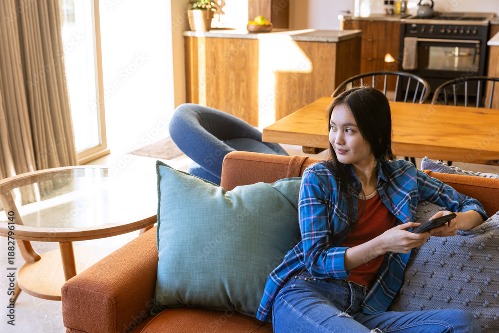 Obraz premium Asian woman sitting on orange sofa holding smartphone in sunlit living area