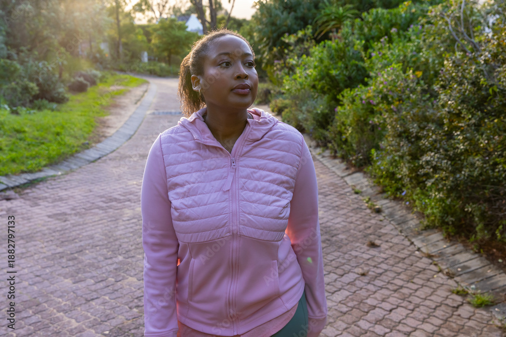 Fototapeta premium African american woman walking through park on brick-paved path wearing jacket, earbuds