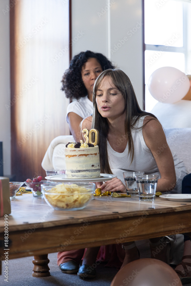 Obraz premium Diverse female friends blowing out 30 candles on cake, balloons in living room