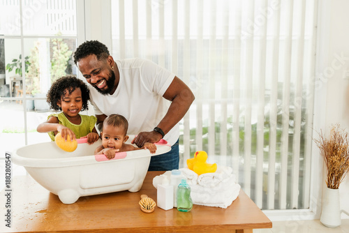 Fotografie Father bathes two young children in a small tub indoors, smiling and playing together in a bright, cozy home environment