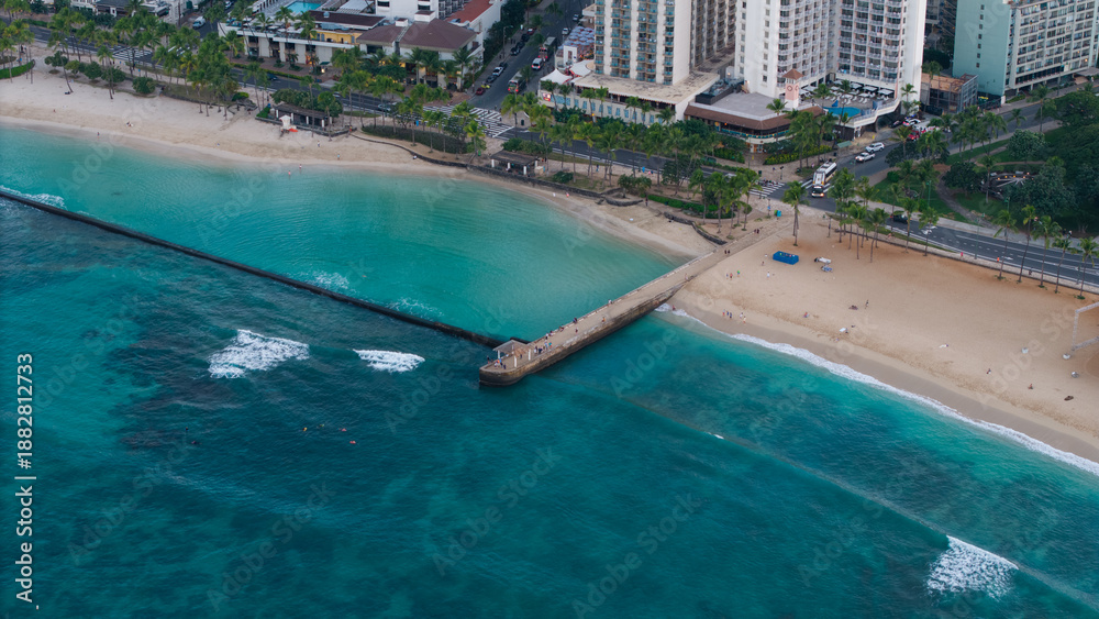 Fototapeta premium Aerial drone view of tropical beach with turquoise ocean water, pier extending into the sea, and coastal city shoreline, scenic island travel destination with clear water and sandy beach