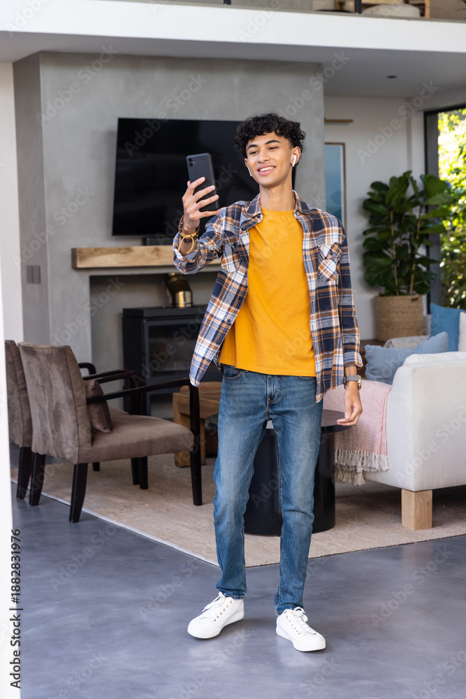 Fototapeta premium Asian male student standing in living room holding smartphone while wearing wireless earbuds