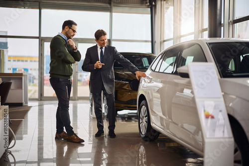 Canvastavla Smiling salesman explaining the features of a new car to his customer in showroom