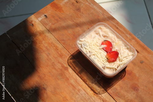 Foto Healthy food for breakfast, a simple fruit salad with lots of cheese toppings and strawberry slices in a plastic box on an aesthetic table bathed in morning sun