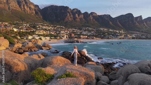 A serene view of Cape Town's coastline at sunset, featuring a woman standing on boulders with Table Mountain in the background