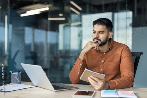 Foto Young man with beard sitting at desk, holding a notebook and pen, looking away f