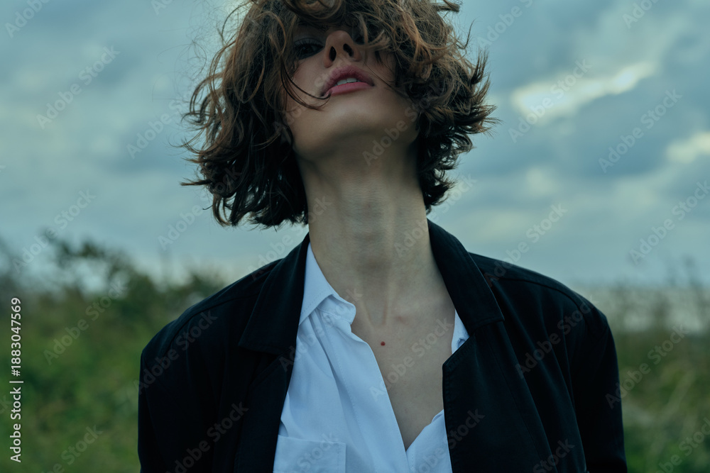 Fototapeta premium Young woman with tousled hair, blazer and white shirt, looking up under a cloudy sky in an open outdoor setting, moody fashion shot