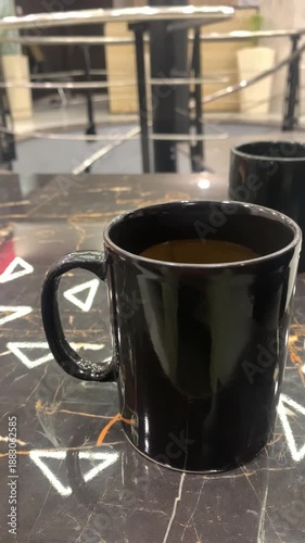 Hot coffee with milk in a glossy black ceramic mug on a dark marble table with geometric patterns.