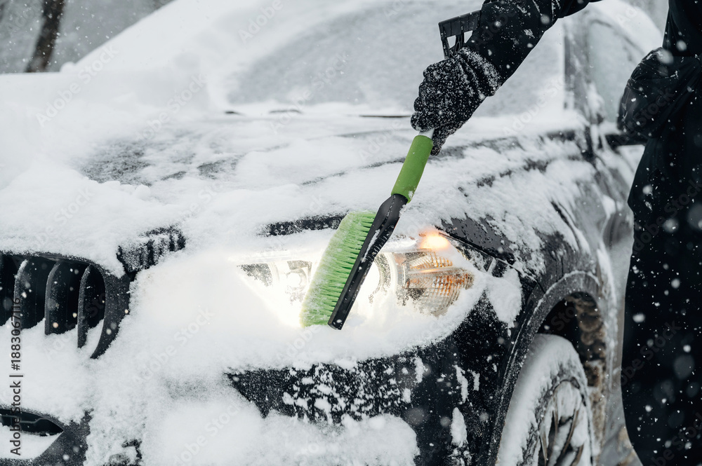 Fototapeta premium Person using a green snow brush to clear snow from the hood and headlights of a black car during a heavy snowfall in a winter environment with falling snowflakes