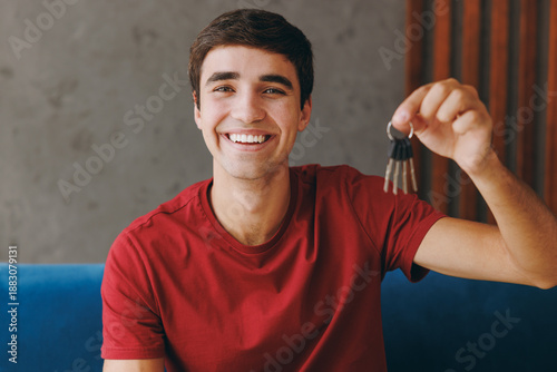 Young happy smiling man he wears red t-shirt hold bunch of keys sit on blue sofa couch stay at home hotel flat rest relax spend free spare time in living room indoors grey wall. People lounge concept.