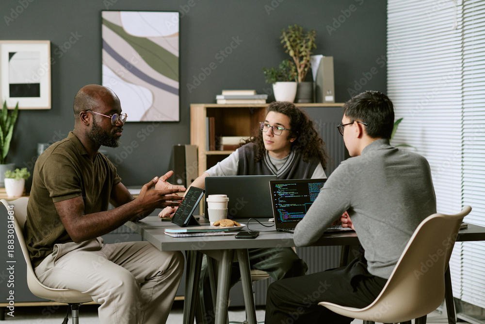 © AnnaStills - Multiethnic group of young adult men and man collaborating around table, discussing project while using laptops with visible code, coffee cup and notepad on tabletop