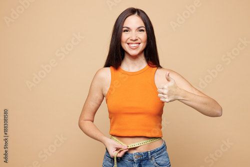 Young smiling woman wear orange casual clothes hold measure tape on waist show thumb up isolated on plain pastel light beige background. Proper nutrition healthy fast food unhealthy choice concept.