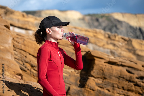 sporty woman wearing red sportswear drinks water from a bottle while exercising outdoors against rugged rocks