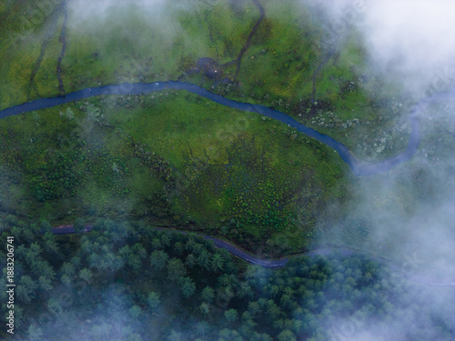 Aerial view of a winding river cutting through the lush green valley, embraced by ethereal clouds and dense forest, Phobjikha Valley, Wangdue Phodrang, Bhutan.