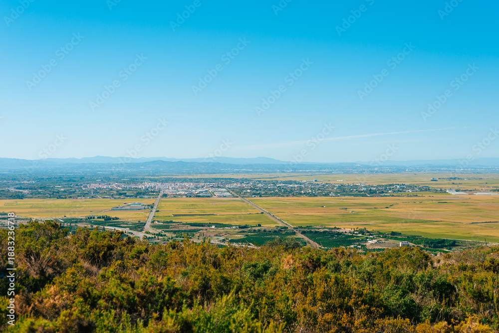 Fototapeta premium A wide view shows a city surrounded by fields and distant mountains. The scene captures a clear day with blue sky. Roads line the fields, connecting the landscape