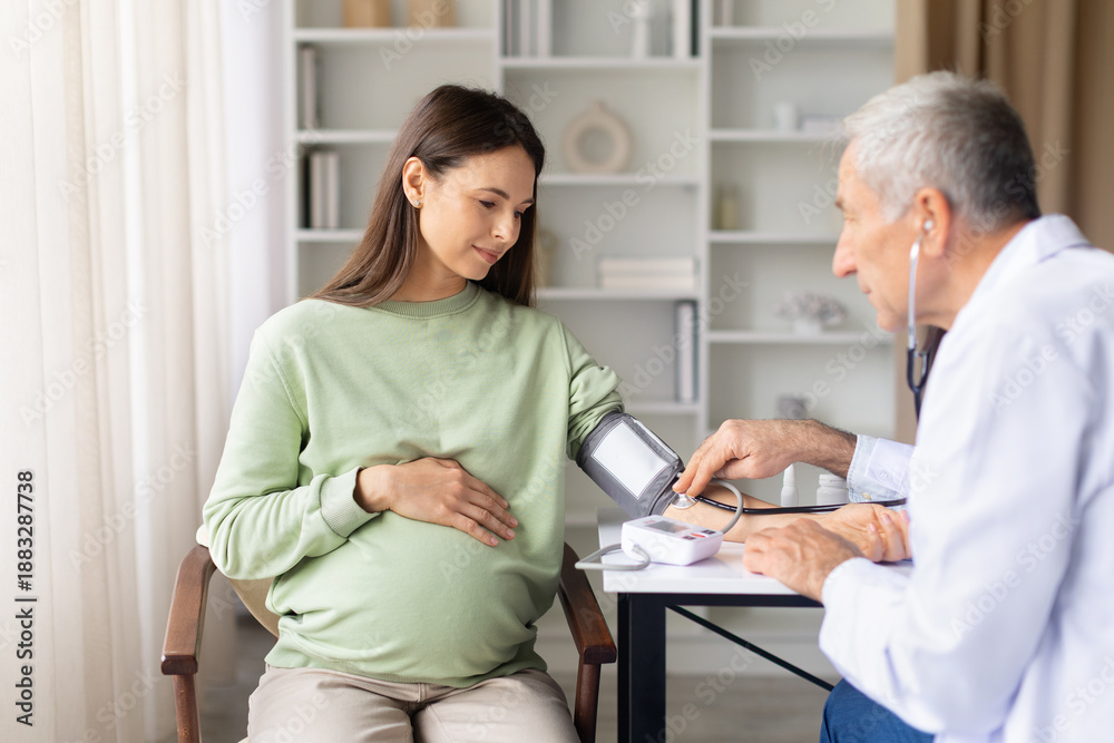 Obraz premium Senior male doctor measuring blood pressure of pregnant woman during prenatal examination, maternity health monitoring and routine pregnancy care