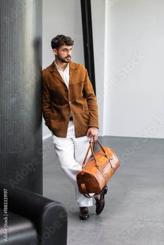 Handsome stylish man in brown suede blazer and white trousers holding premium leather duffel bag in modern interior