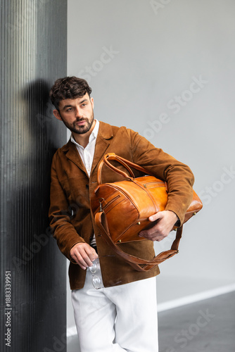 Fashionable man in a brown suede blazer and white trousers leaning against a wall while holding a leather travel bag