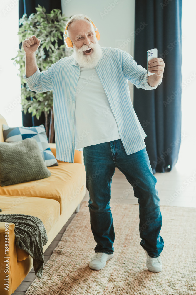 Fototapeta premium Senior man with headphones dances in the living room during a joyful moment captured at home on a sunny day