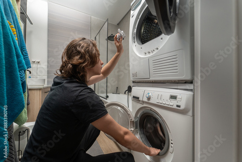 Young man in black t-shirt moving wet laundry from washer to dryer in bathroom. Male person doing laundry chores using stacked home appliances for washing and drying routine at home.